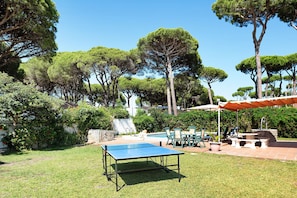 A pool table in a grassy outdoor area with trees and outdoor furniture.