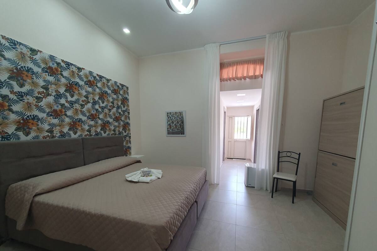 A bedroom with a bed, a nightstand, a chair, a wardrobe, and a door leading to another room.