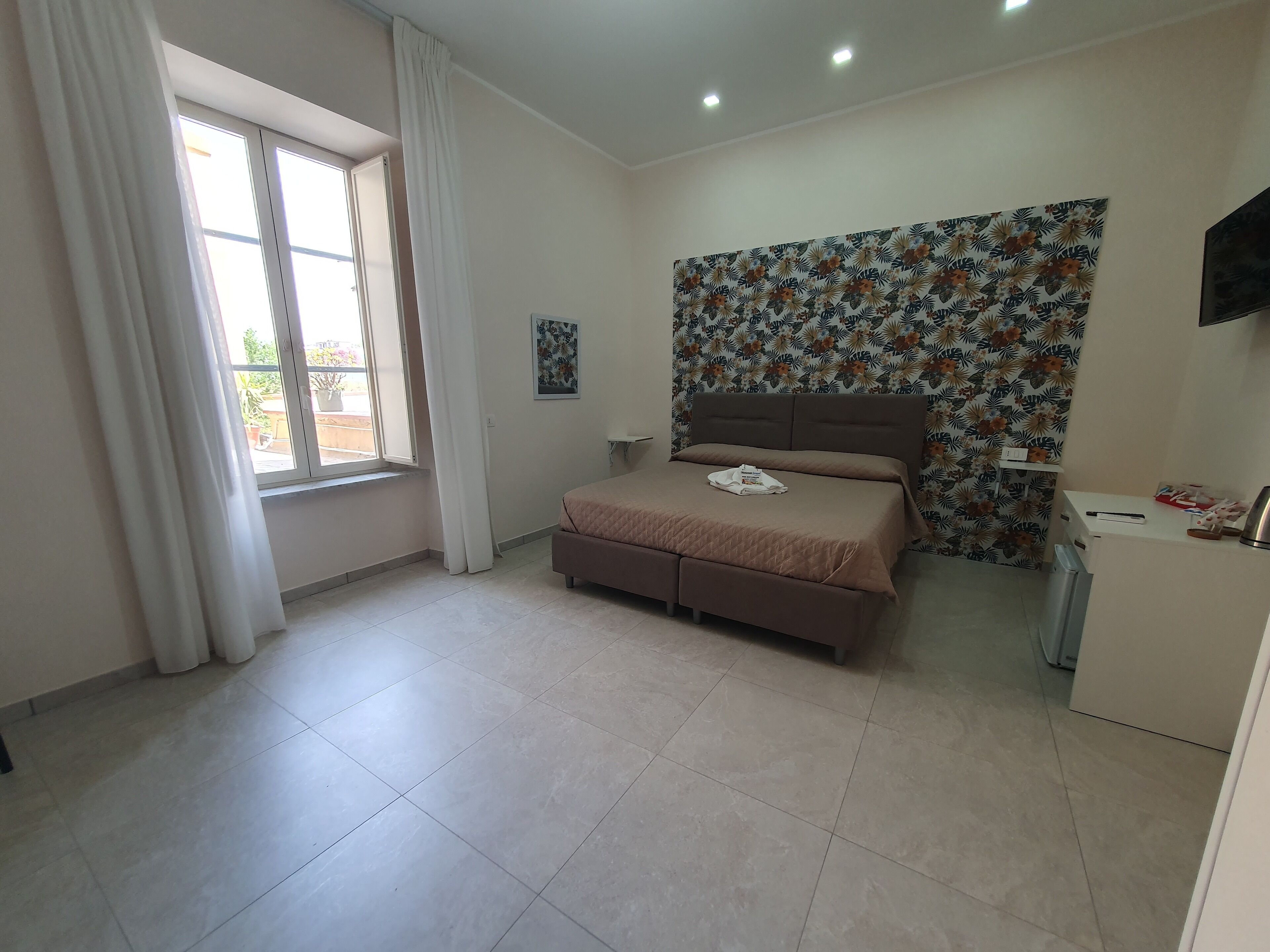 Ein Schlafzimmer mit einem Bett, einem Fenster mit Vorhängen, einer Wand mit Blumenmuster und einem kleinen Schreibtisch mit Wasserkocher.