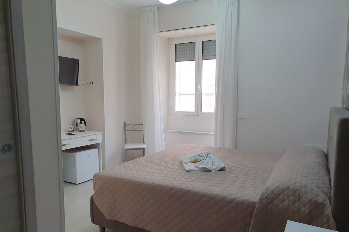 A bedroom with a bed, a desk with a chair, a television on the wall, and a window with curtains.