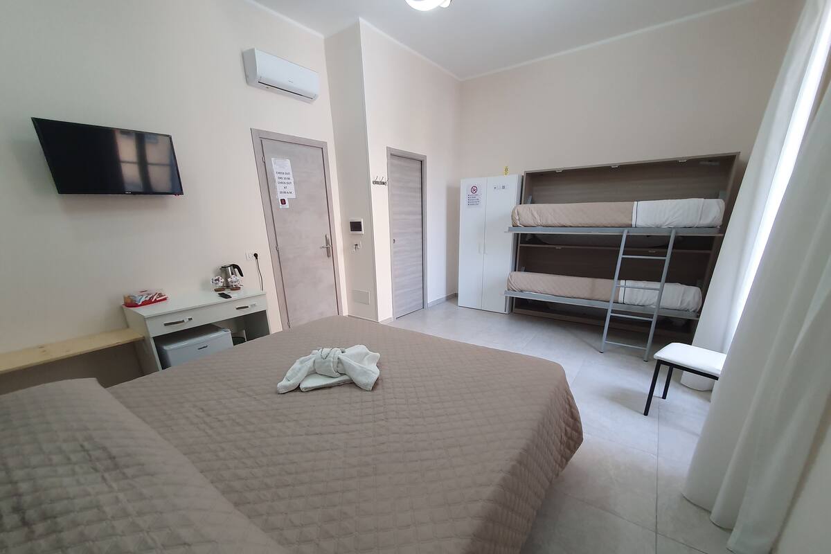 A hotel room with a bed, a desk with a chair, a TV, a wardrobe, and a bunk bed.