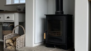 A modern living room with a black wood-burning stove, a stone hearth, a wicker basket with firewood, and a view of the outside through a window with striped valance.