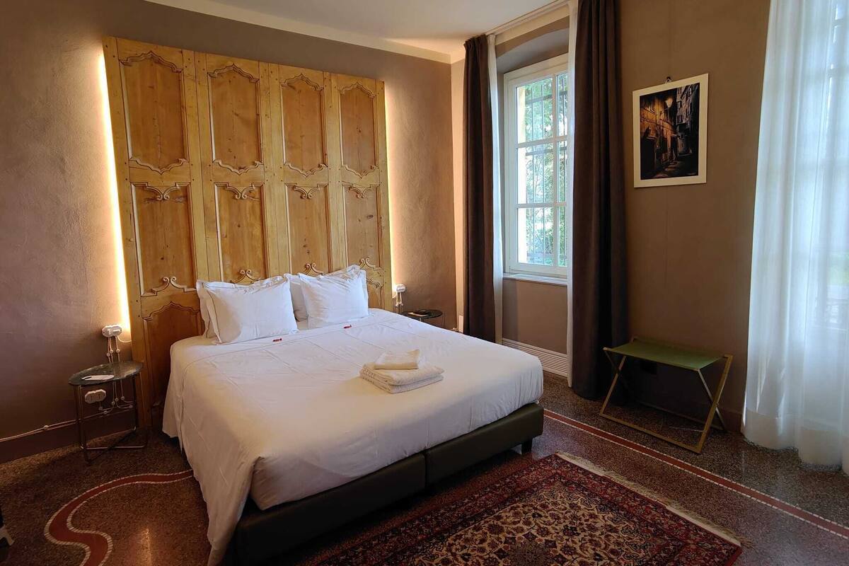 Ein Hotelzimmer mit einem großen Bett, einem hölzernen Kopfteil, einem Fenster mit Vorhängen, einem kleinen Tisch und einem Teppich.