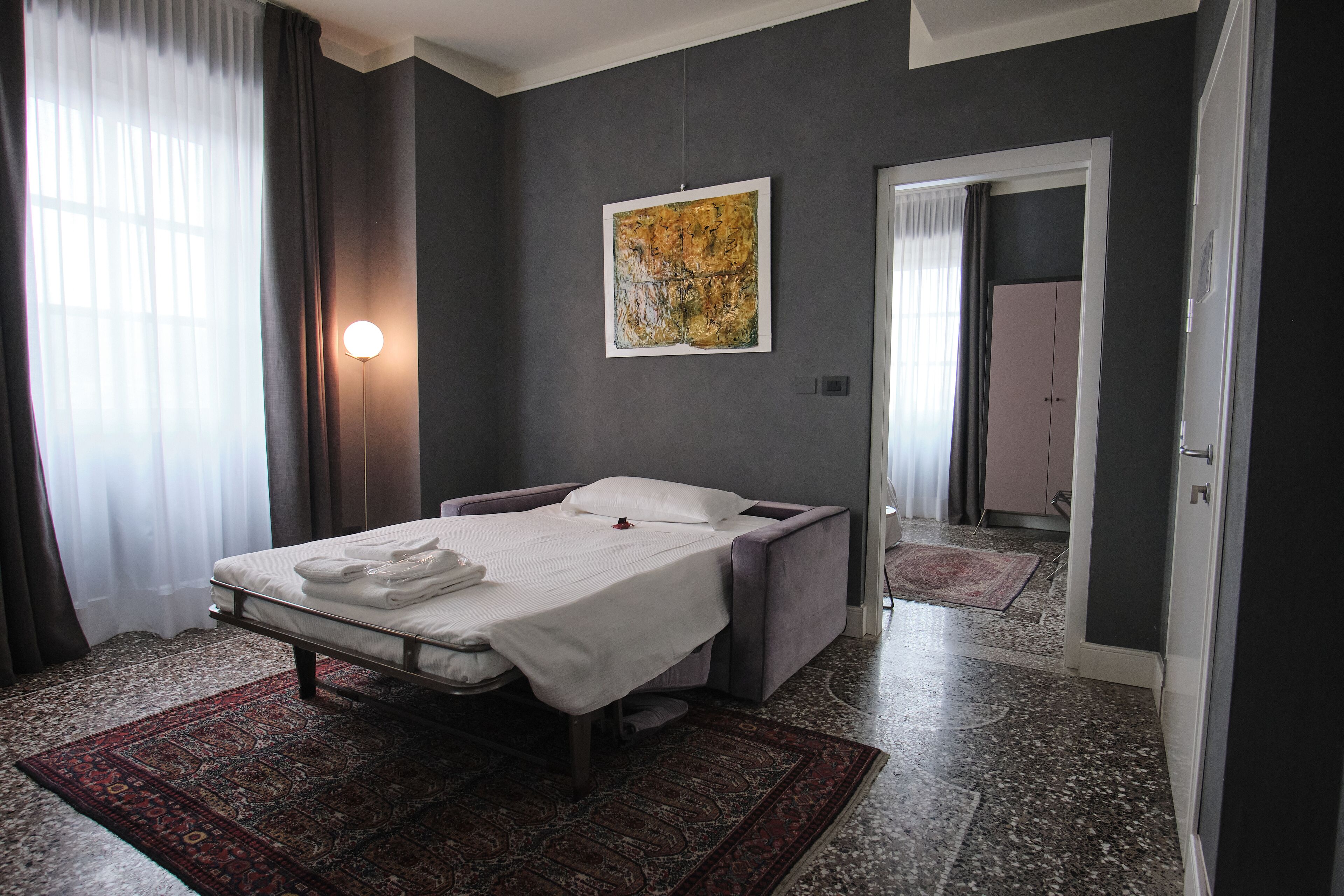A modern hotel room with a bed, a large abstract painting, a standing lamp, and a view of the hallway.