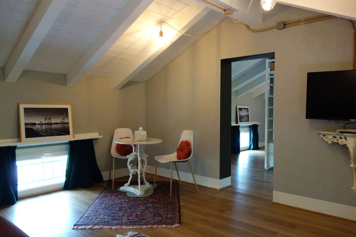 Ein modernes Wohnzimmer mit Holzboden, einem weißen Tisch mit zwei Stühlen, einem gemusterten Teppich, einem gerahmten Bild an der Wand und einem an der Wand montierten Fernseher.
