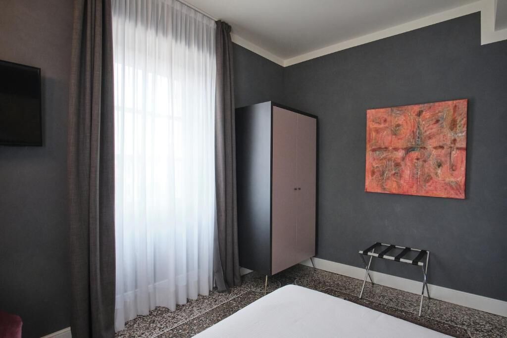 A modern hotel room with a large bed, a wardrobe, a painting on the wall, and a window with curtains.