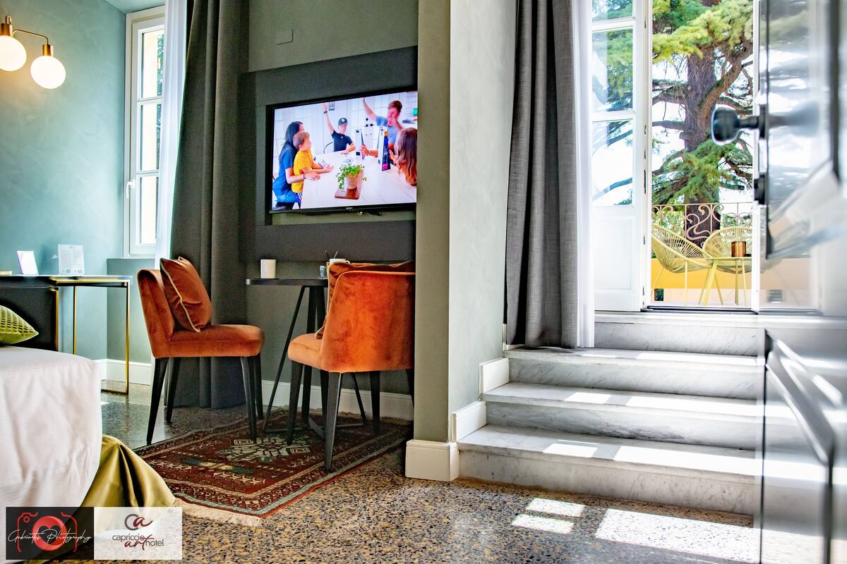 Ein Hotelzimmer mit Fernseher, zwei Stühlen, einem Bett, einem Schreibtisch und Blick auf einen Baum.