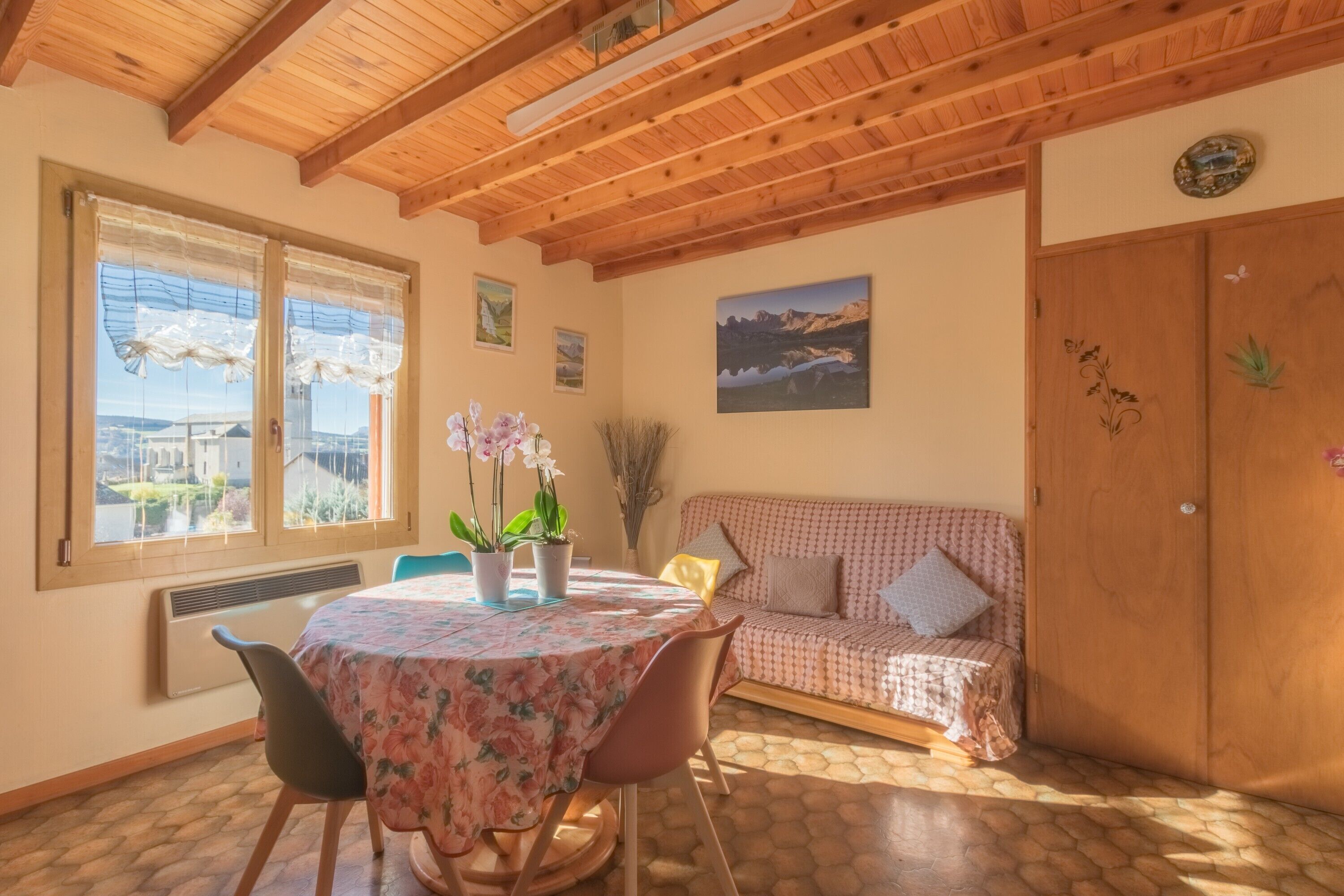 Een knus interieur met een houten plafond, een ronde tafel met bloemendecoratie, een bank en een groot raam met gordijnen.