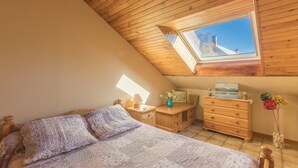 Une chambre avec un plafond en pente, un puits de lumière, un lit recouvert d’une couverture à motifs, une commode en bois et une table de chevet avec une lampe.