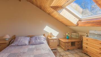 Ein Schlafzimmer mit schrägen Wänden, einem Dachfenster, einem Bett mit gemustertem Überwurf, einem Nachttisch mit Lampe, einem Schminktisch und einem Schreibtisch mit Lampe.