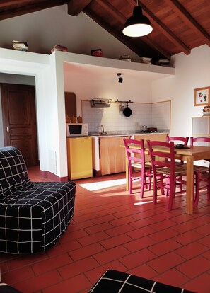 Un salón acogedor con un sofá a cuadros blanco y negro, una mesa de comedor de madera con sillas rosas y una cocina con microondas, hervidor y perchero para ollas.
