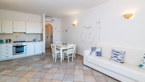 Une cuisine avec des meubles blancs, une table à manger avec des chaises, un canapé blanc avec des coussins à motifs et un mur agrémenté de luminaires décoratifs.