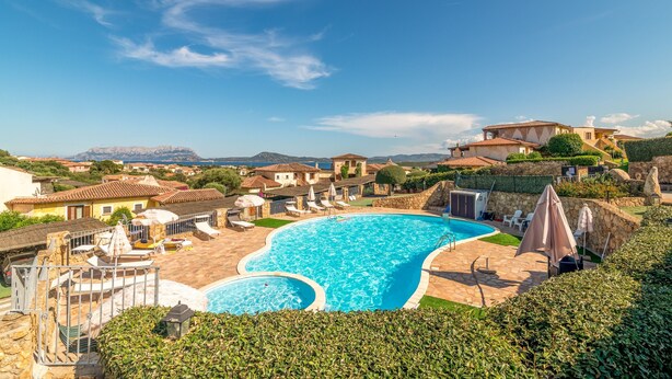 Une grande piscine avec une terrasse en pierre, entourée de maisons à l’architecture méditerranéenne, et un ciel dégagé.