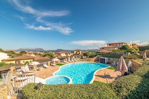 Une grande piscine avec une terrasse en pierre, entourée de maisons à l’architecture méditerranéenne, et un ciel dégagé.