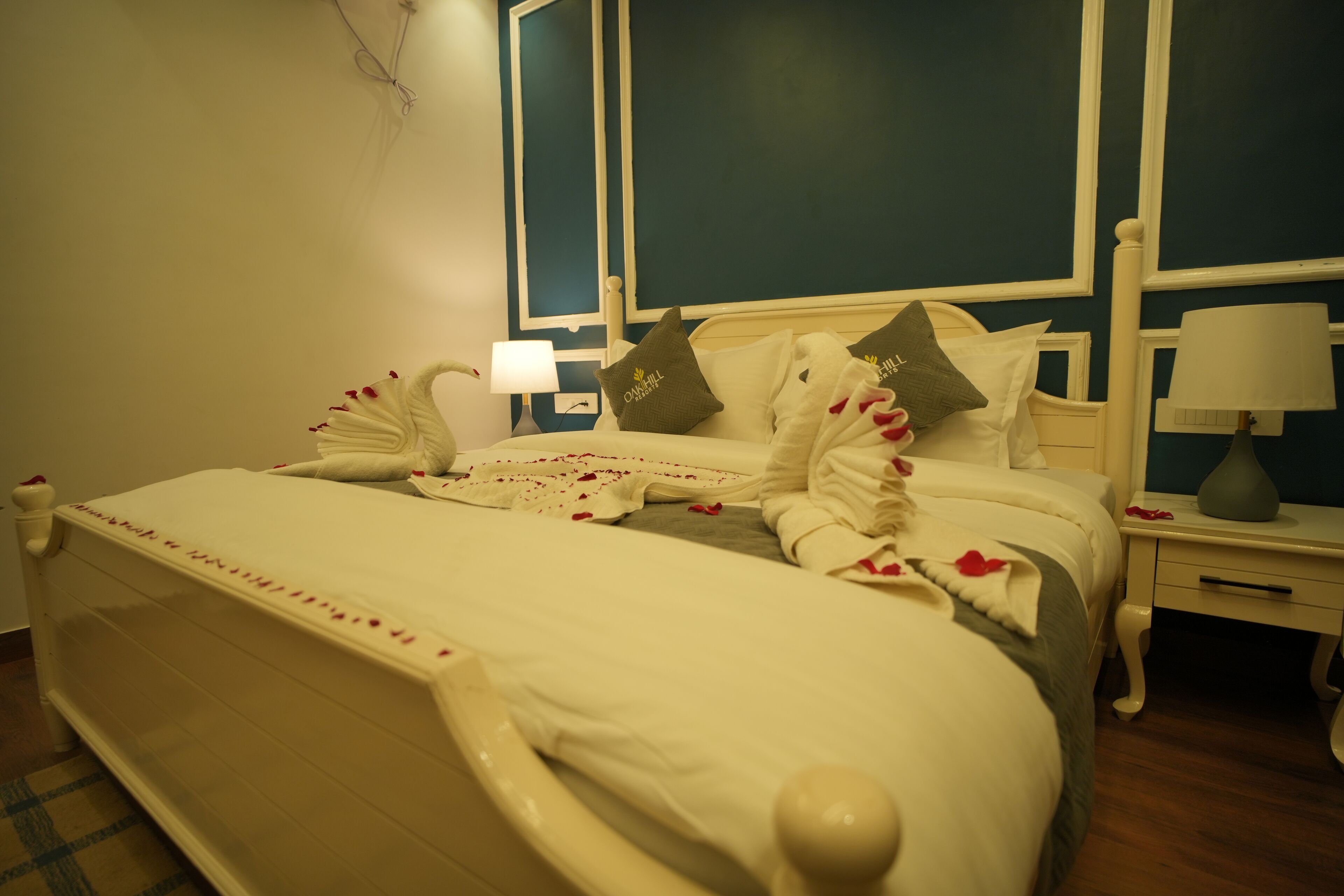 A neatly made bed with a white comforter and pillows, a bedside table with a lamp, and a wall with a mirror and a picture frame.