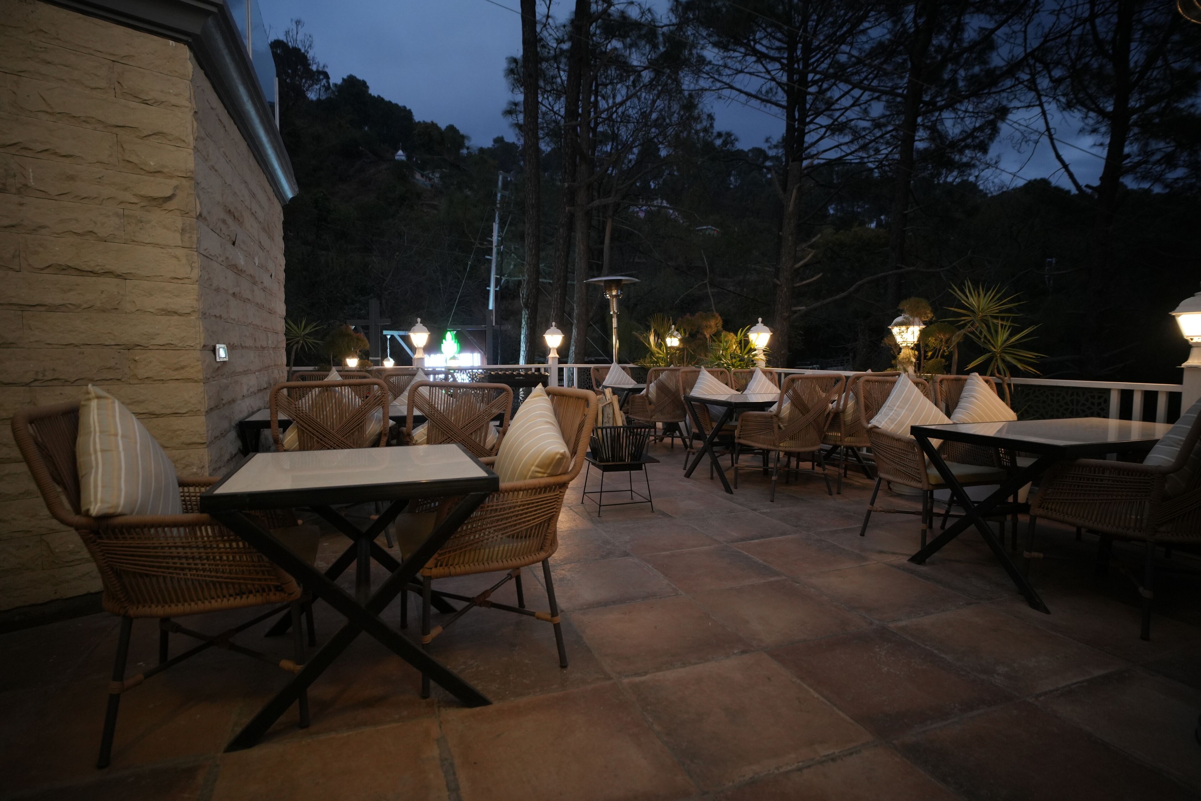 An outdoor patio area with wicker furniture and tables, surrounded by trees and a stone wall.