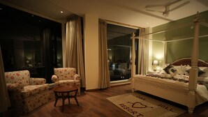 A bedroom with a four-poster bed, a sitting area with floral armchairs, a small round table, and a view of the outside through a large window. - Oakhill Resort (Kasauli)