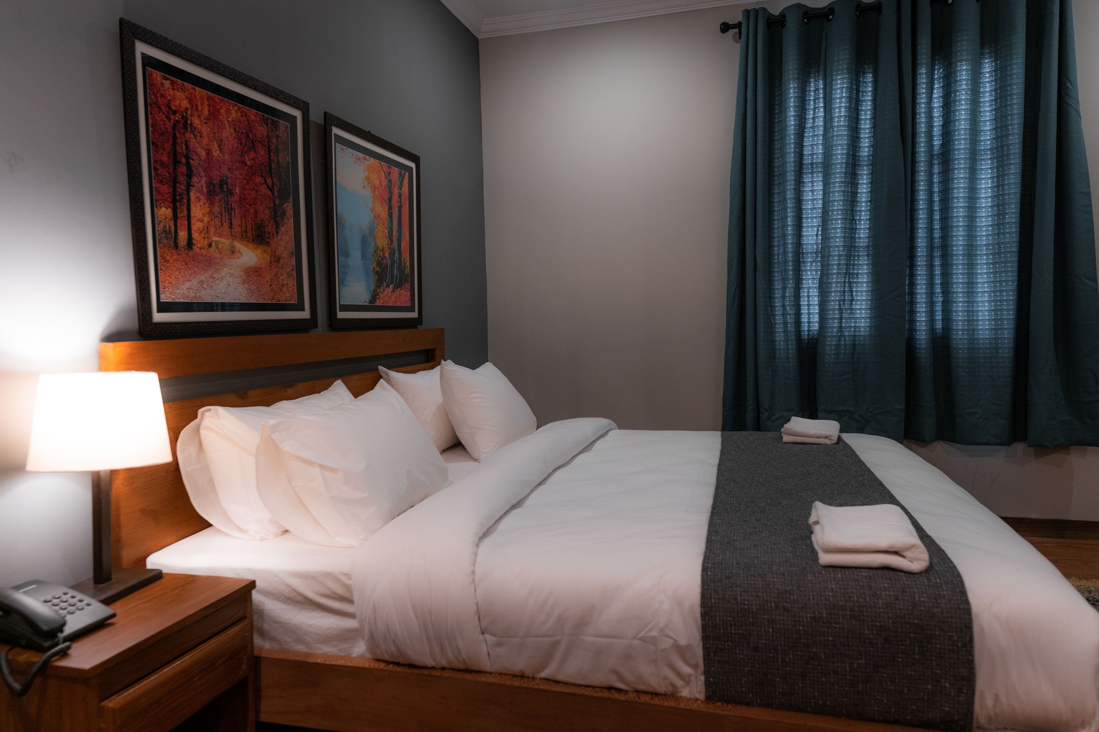 Una cama bien hecha con ropa blanca, una lámpara de noche y un teléfono sobre una mesita de madera. Dos cuadros enmarcados cuelgan sobre la cama.