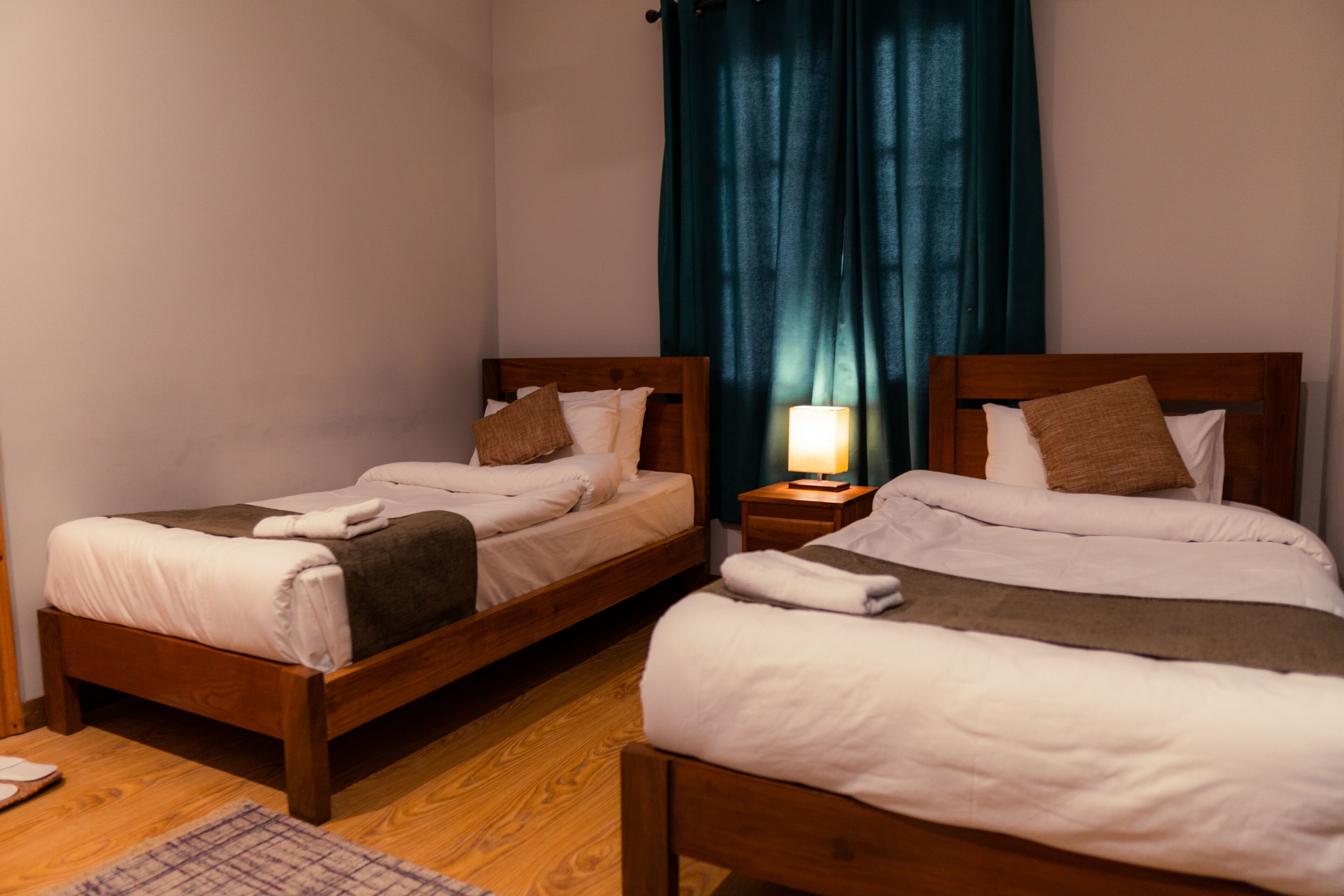 Una habitación con dos camas de madera, cada una con ropa blanca y almohadas marrones. Hay una mesita de noche pequeña con una lámpara entre las camas.