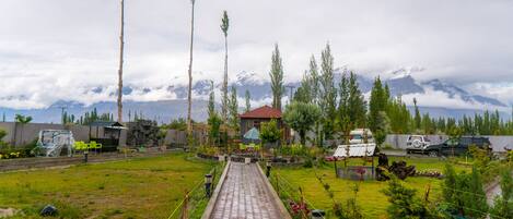 A paved pathway leading to a house with a red roof, surrounded by greenery and a mountainous backdrop.