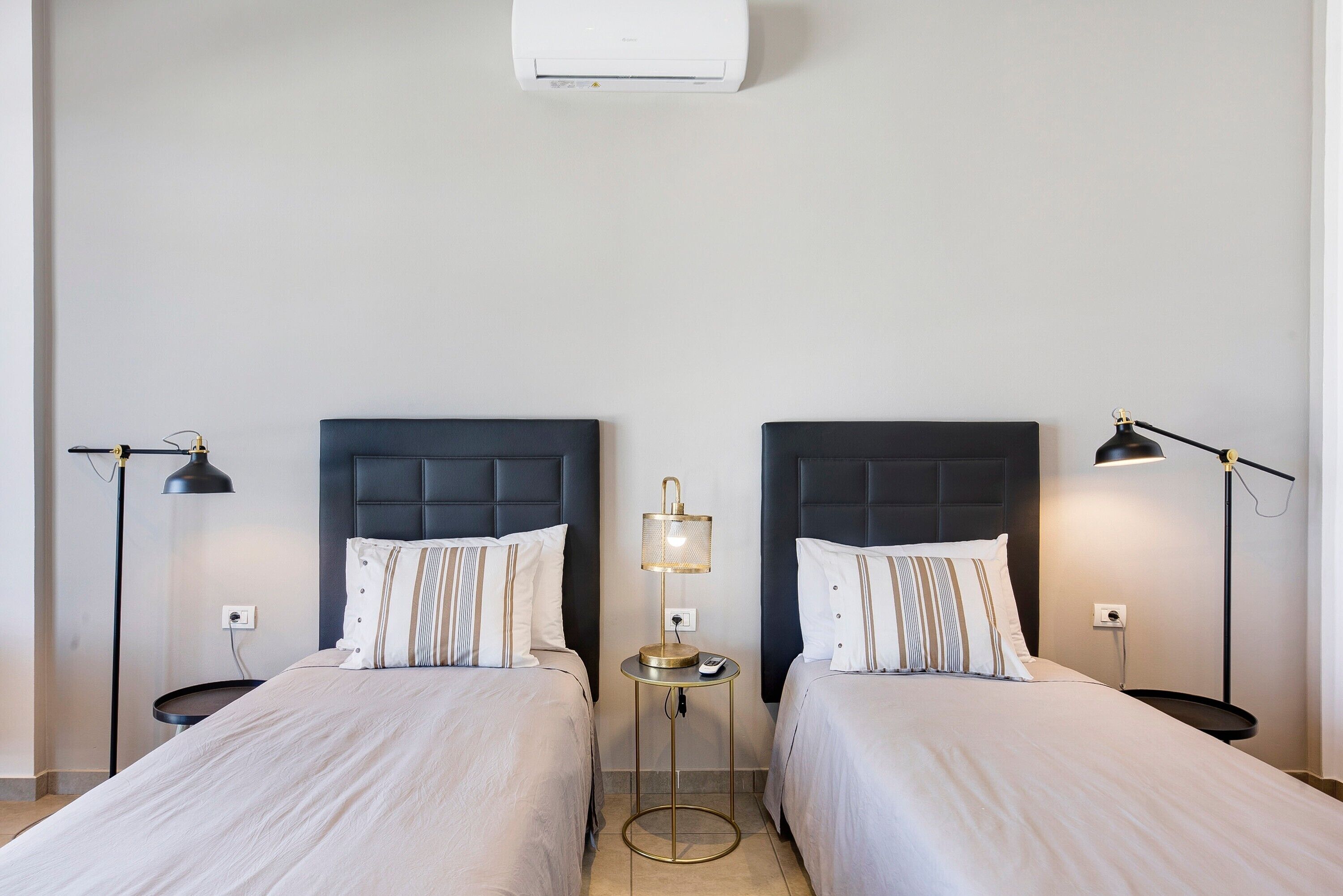 A hotel room with two beds, bedside lamps, a small table, and an air conditioning unit.