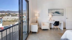 Una habitación de hotel con balcón, escritorio, dos sillas, un cuadro y una cama.