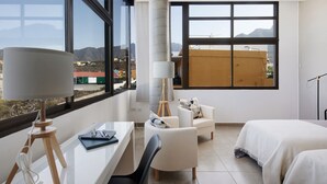 A modern hotel room with large windows, a desk, a chair, a bed, and a lamp.