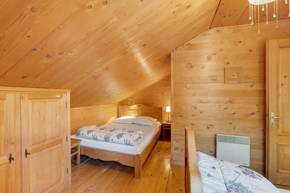 Une pièce en bois avec un plafond en pente, un lit recouvert d’une literie à motifs floraux, une table de chevet avec une lampe et un radiateur.