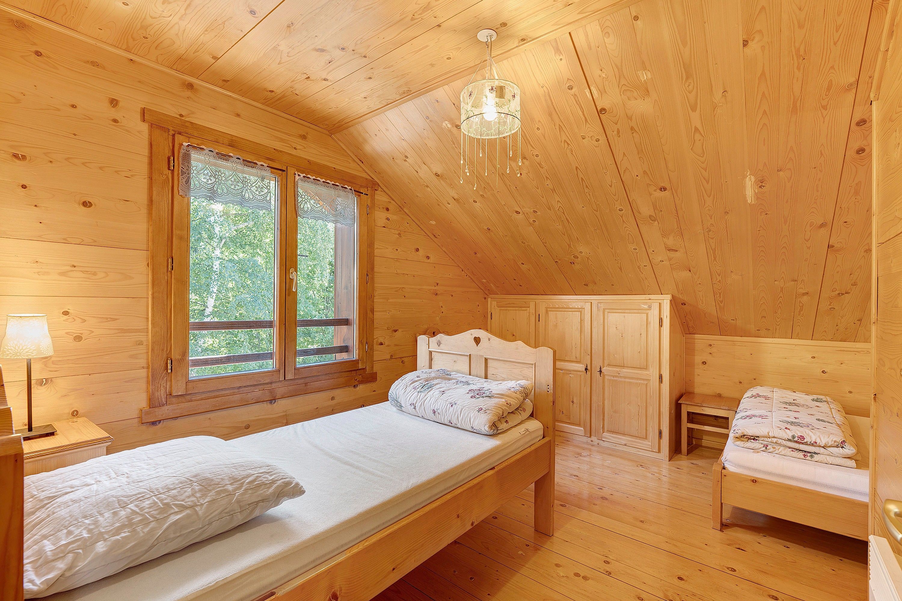Quarto de cabana de madeira com uma cama de solteiro, mesa de cabeceira, cômoda e janela com cortinas de renda.