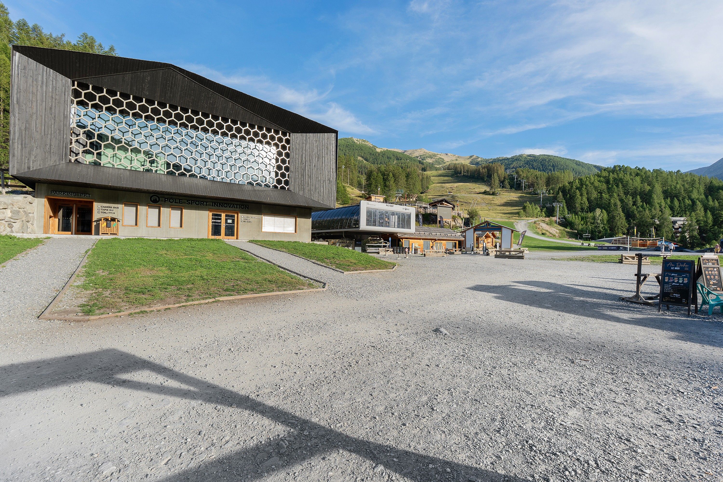 Un bâtiment moderne doté d’une façade en verre hexagonale, d’un parking en gravier et surplombant un paysage montagneux.