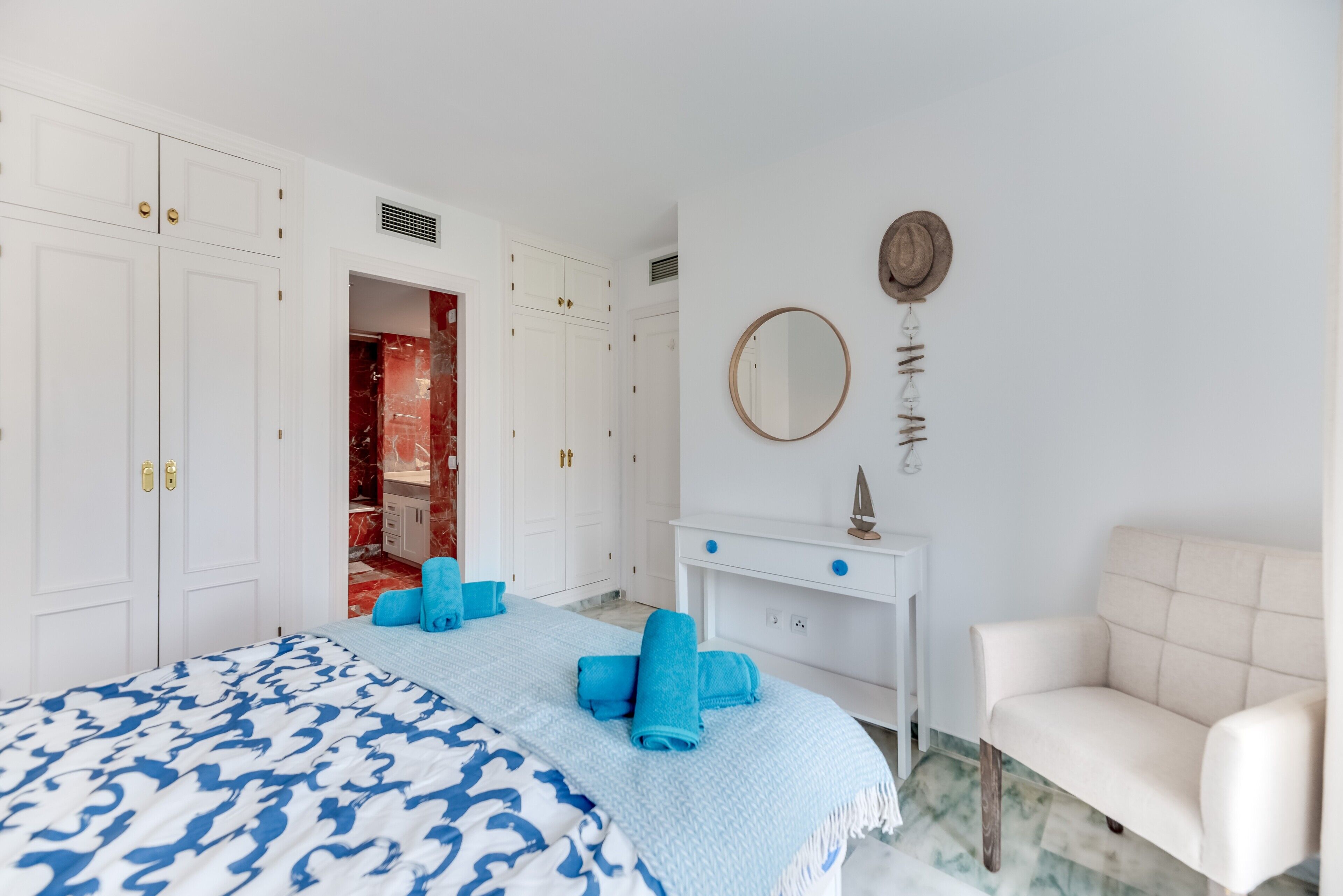 A bedroom with a bed, white wardrobe, a vanity with a mirror, and a white armchair.
