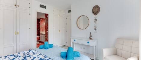 A bedroom with a bed, white wardrobe, a vanity with a mirror, and a white armchair.