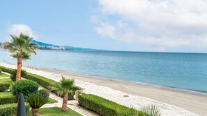 Una playa con vista despejada al mar, un camino bordeado de palmeras y una cadena montañosa distante.