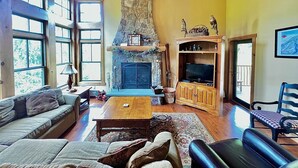 Un salón amplio con chimenea de piedra, mobiliario de madera y ventanas grandes.