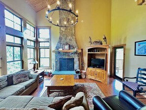 A spacious living room with a stone fireplace, wooden furniture, and large windows.