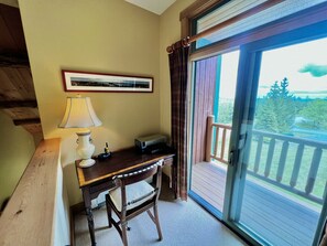 A room with a desk, chair, lamp, and a large window with a view of trees and a deck.