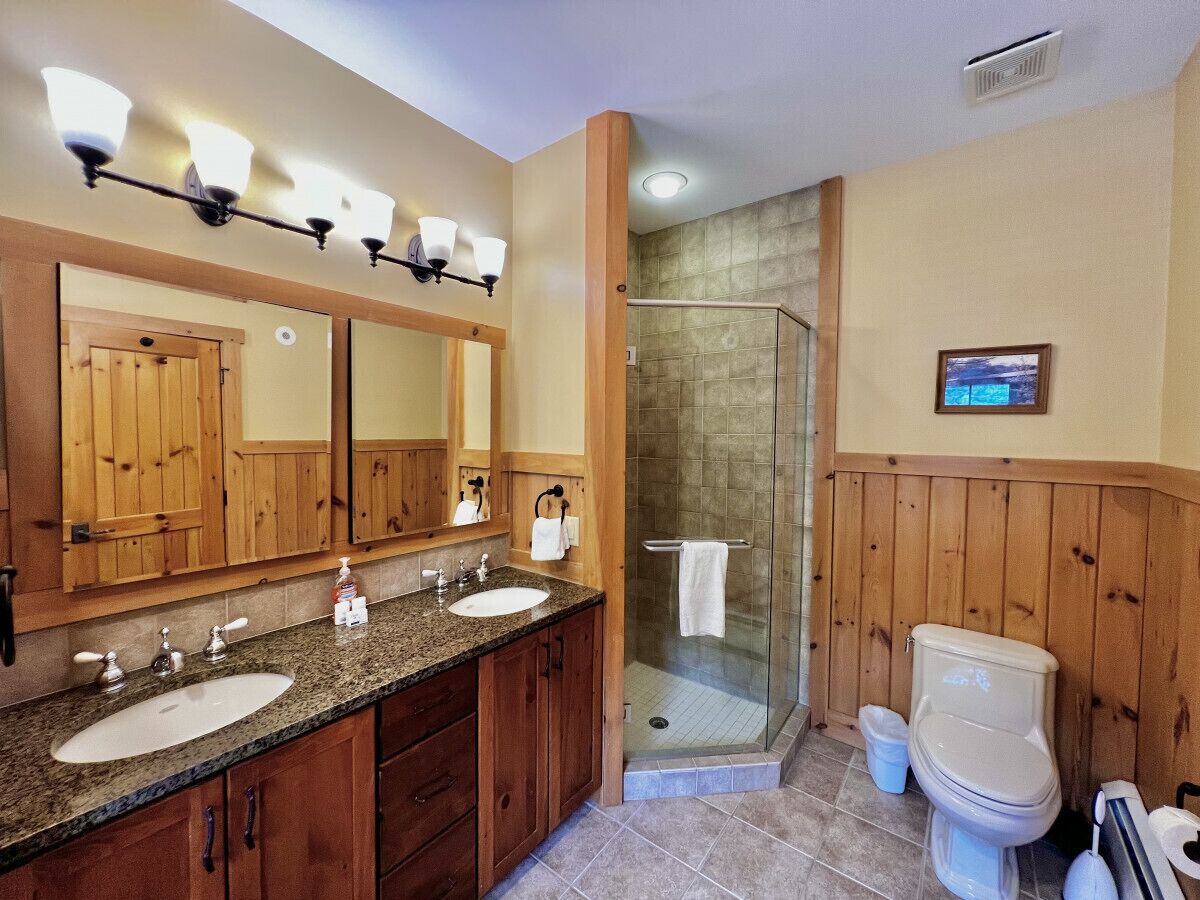 Een badkamer met houten lambrisering, een dubbele wastafel, een douchecabine van glas en een toilet.