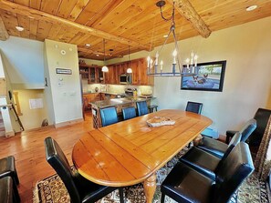A spacious dining area with a large wooden table and black chairs, a kitchen in the background, and a chandelier above.