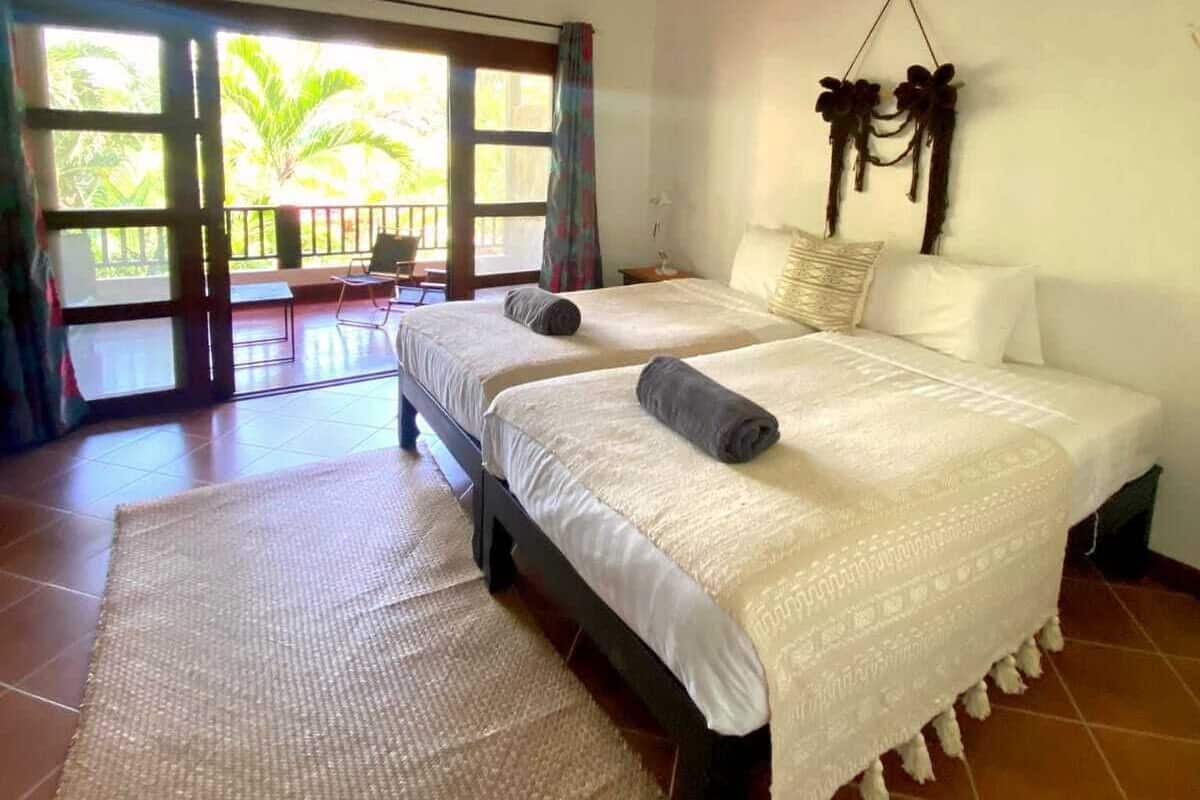Quarto de hotel com duas camas, ventoinha no teto, uma janela grande com cortinas e vista para o exterior.