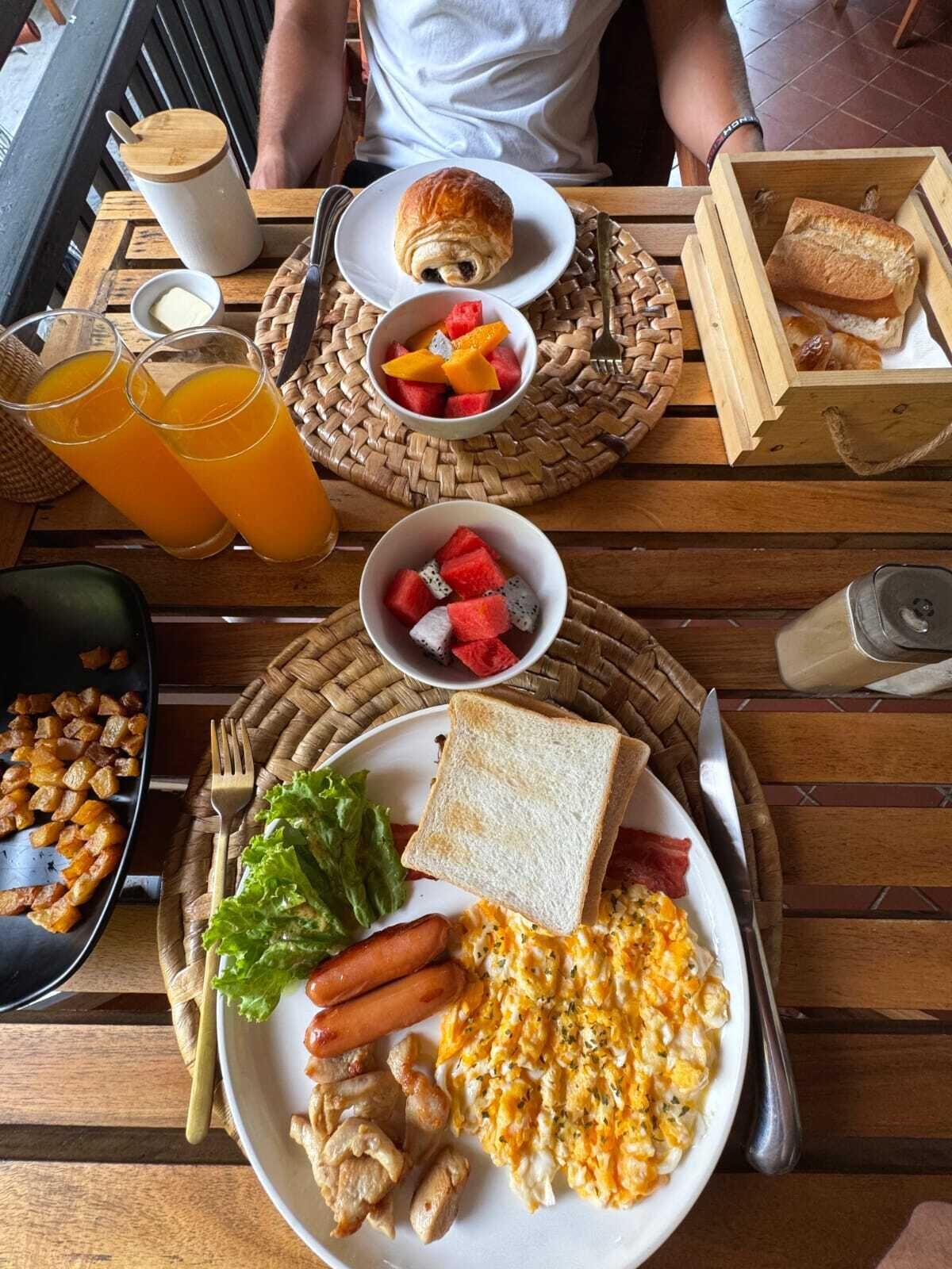 A breakfast setting with a plate of scrambled eggs, sausage, and bacon, alongside toast, fruit, and a croissant.