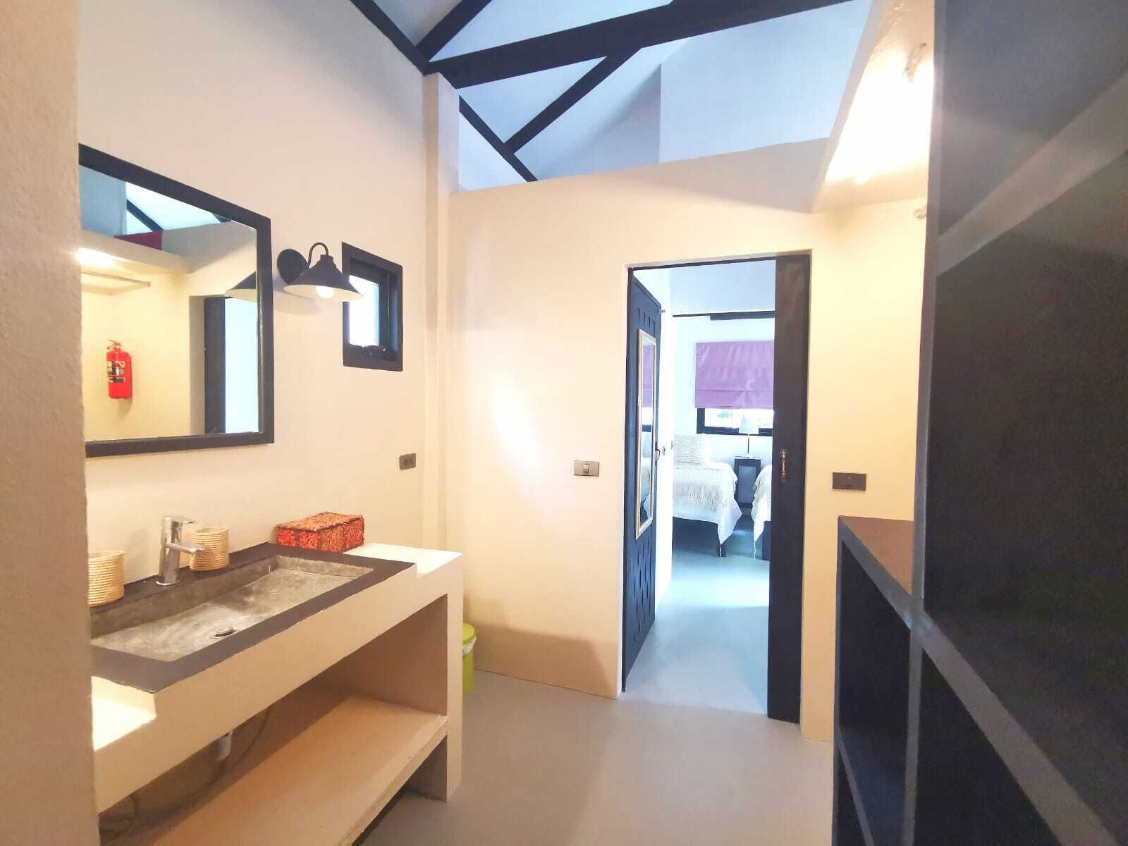 A modern bathroom with a large mirror, a sink, and a toilet. There is a hallway leading to a bedroom with a bed and a window.