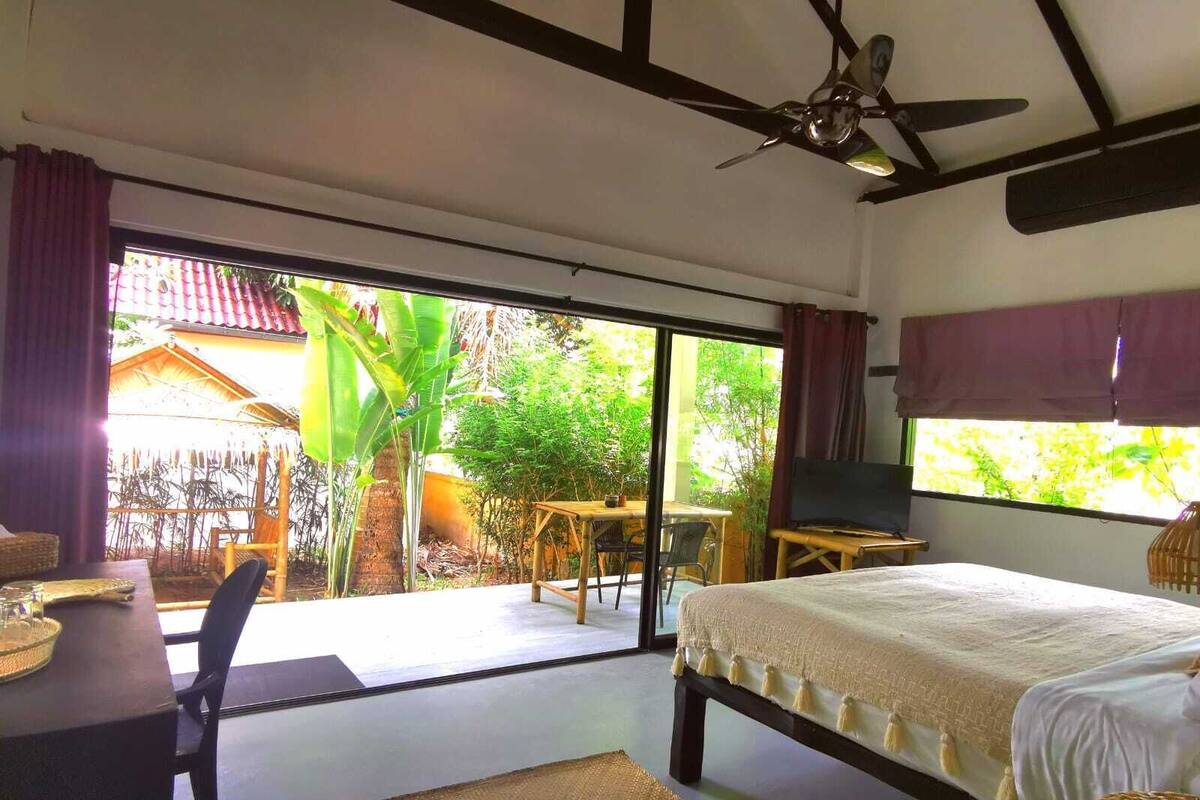 Quarto com cama, ventilador de teto, televisão e vista para um jardim através de amplas portas de correr.