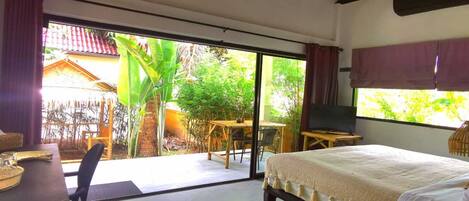 Un dormitorio con cama, ventilador de techo, televisor y vistas a un jardín a través de amplios ventanales correderas.