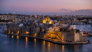 Paisatge urbà costaner a la penumbra, amb edificis històrics, un port fortificat i una església destacada.