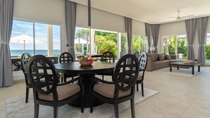 Uma sala de estar ampla com uma mesa de jantar cercada por cadeiras, um sofá e uma mesa de centro. Grandes janelas oferecem vista para o oceano e vegetação exuberante.