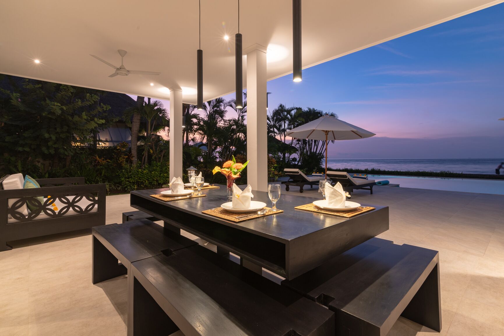 Un área de comedor al aire libre con una mesa preparada para cuatro personas, con vistas a una piscina y al océano al atardecer.