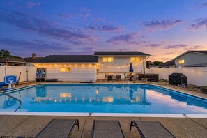 A rectangular swimming pool with a 5 ft depth marker, surrounded by lounge chairs, a barbecue grill, and string lights. - Modern 3BR w/ Pool, Hot Tub & Fenced Yard (Parma)