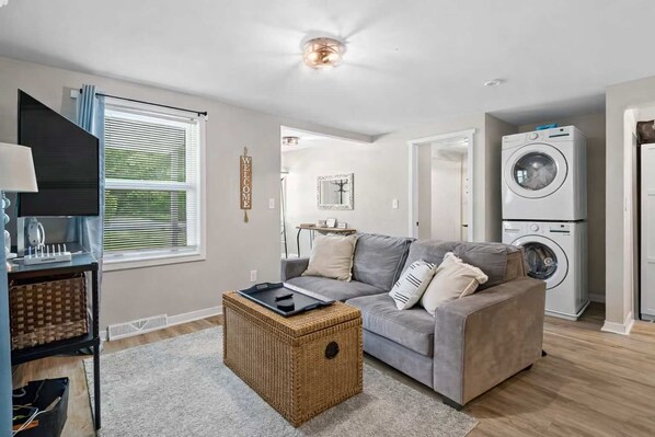 A modern living room with a grey sofa, a wicker coffee table, a TV, and a washing machine.