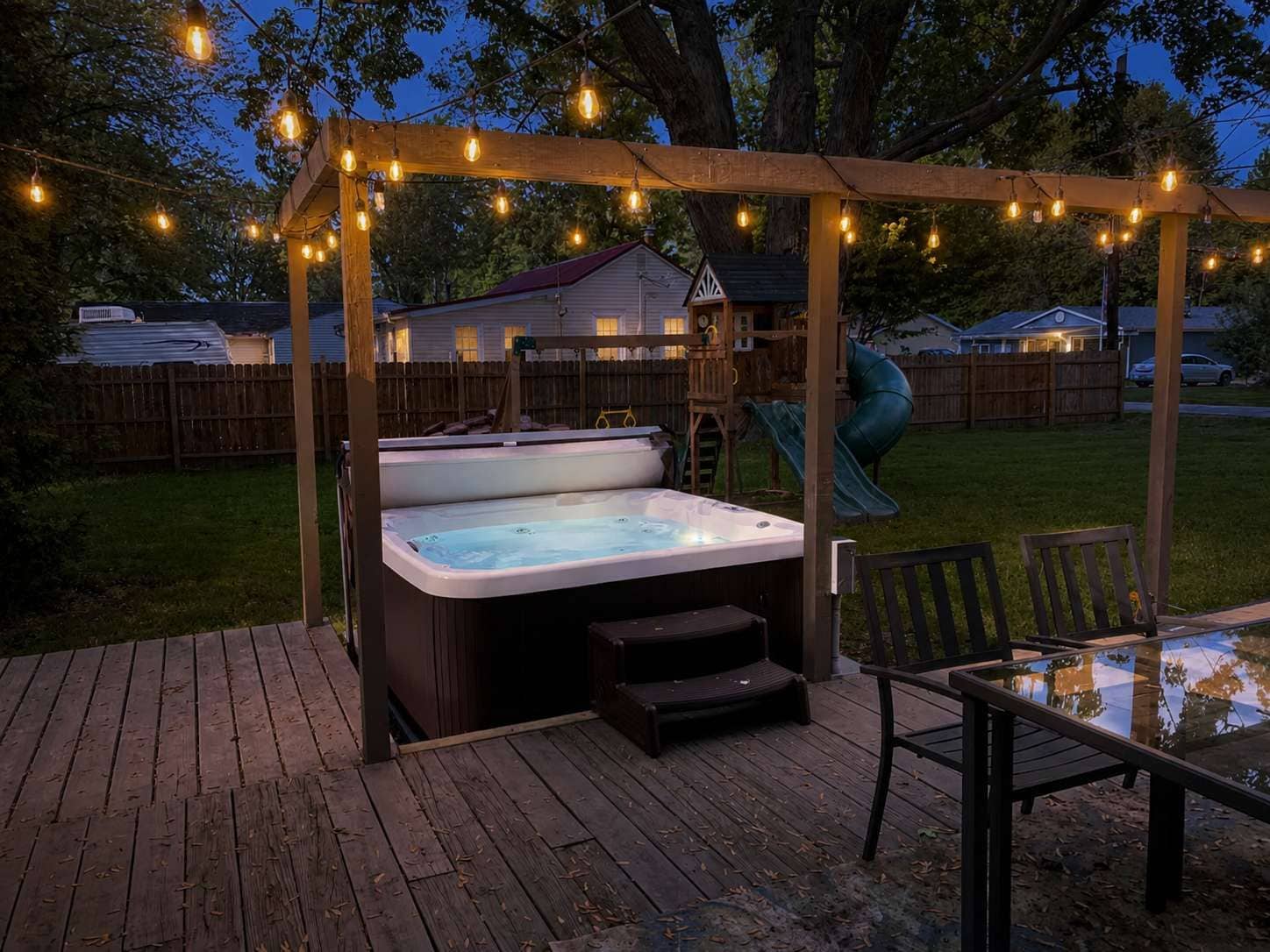 A hot tub under a wooden pergola with string lights, a slide, and a deck with chairs.