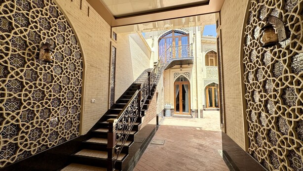 Un couloir aux murs ornés de motifs géométriques complexes, un escalier avec des rampes richement décorées, et une vue sur un bâtiment aux éléments architecturaux soignés.